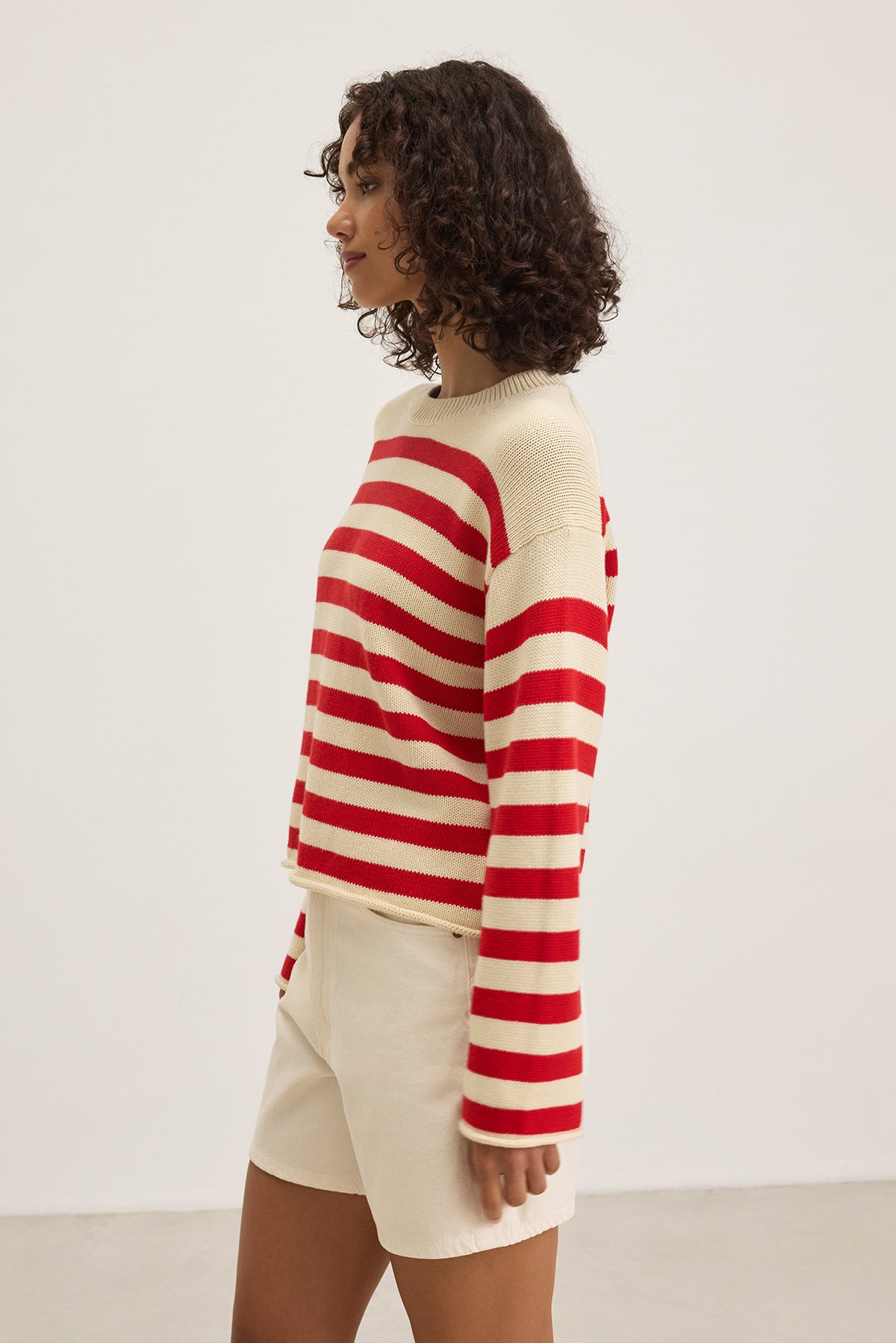 A person with curly hair is standing in profile against a plain background, wearing a LEX COTTON CASHMERE SWEATER by Velvet by Graham & Spencer in a red and white Breton stripe pattern, complemented by beige shorts.-38662726516929