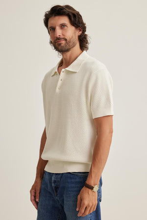 A bearded man in a Velvet by Graham & Spencer LOWELL Cotton Cashmere Mesh Sweater Polo and blue jeans stands against a plain background.