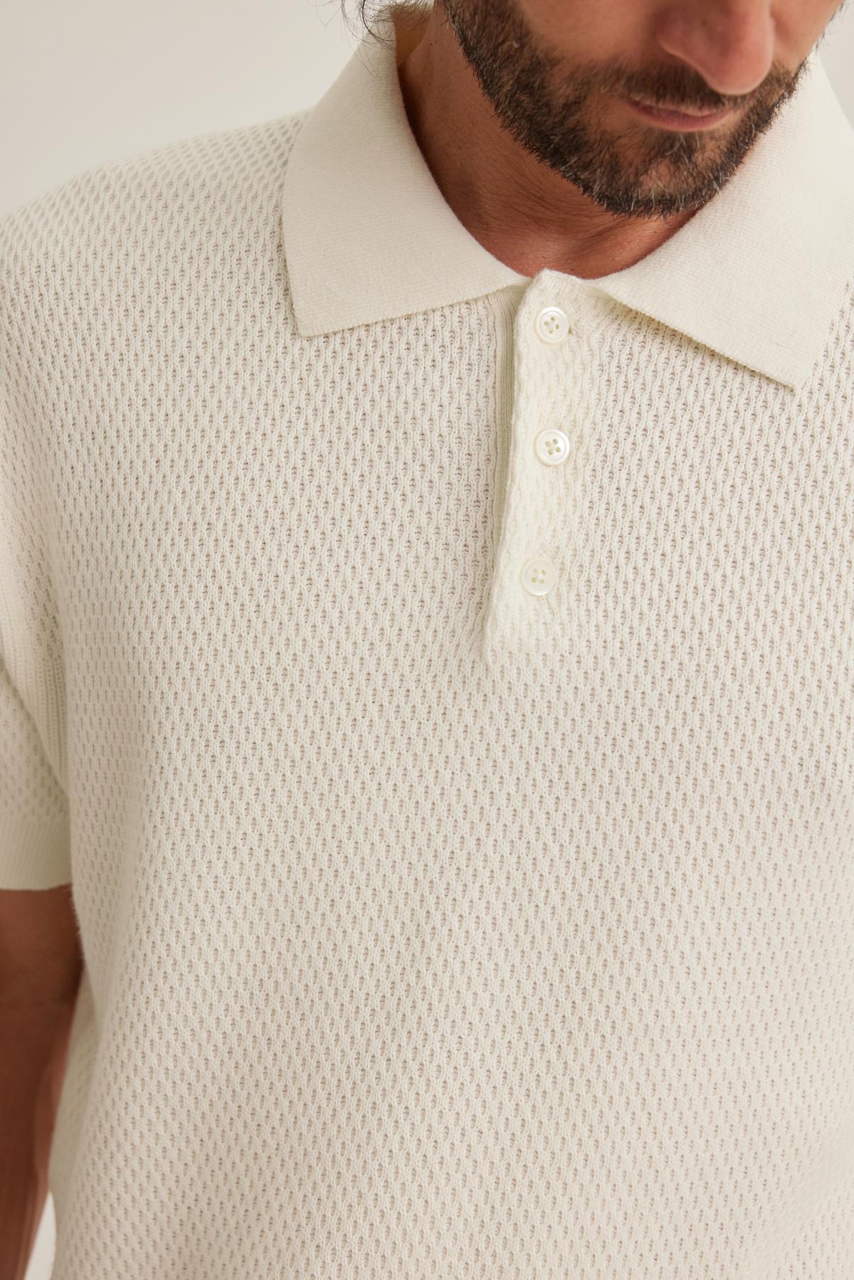 A man wearing a Lowell Cotton Cashmere Mesh Sweater Polo by Velvet by Graham & Spencer, featuring a textured white design with a collar and three buttons, is looking downwards.