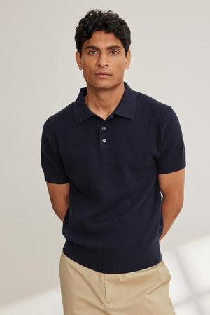 A person in a LOWELL Cotton Cashmere Mesh Sweater Polo by Velvet by Graham & Spencer and beige pants stands with hands behind their back against a neutral background.