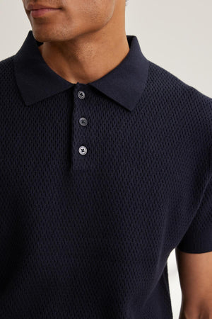 A person wearing the Velvet by Graham & Spencer Lowell Cotton Cashmere Mesh Sweater Polo, featuring a textured, short-sleeve design in black with three buttons. The focus is on the upper torso and shirt details.