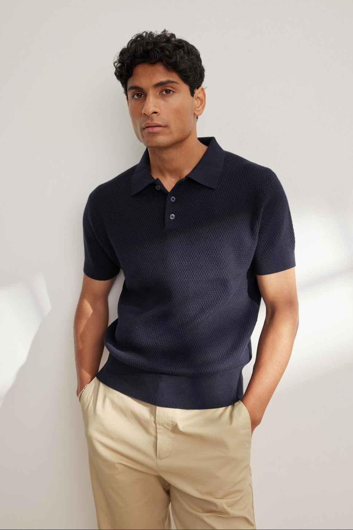 A man stands against a white wall wearing the LOWELL SWEATER POLO by Velvet by Graham & Spencer in navy with beige pants—hands in pockets—a versatile staple for any wardrobe.-38643405390017