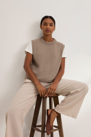 A person sits on a wooden stool, wearing a beige sleeveless sweater over a white top, the KELLY PANT—wide-leg striped pants with an elastic waist—from Velvet by Graham & Spencer, and brown shoes, set against a plain white background.