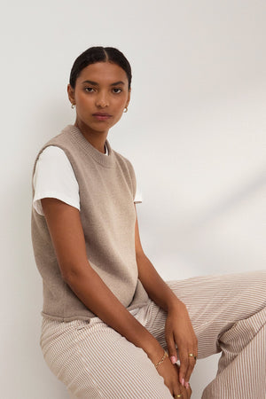 A person sits against a white wall wearing Velvet by Graham & Spencer's PAOLA COTTON CASHMERE SWEATER VEST over a white T-shirt and striped pants. Their short hair frames their face as they look at the camera, giving off a casual yet polished vibe.
