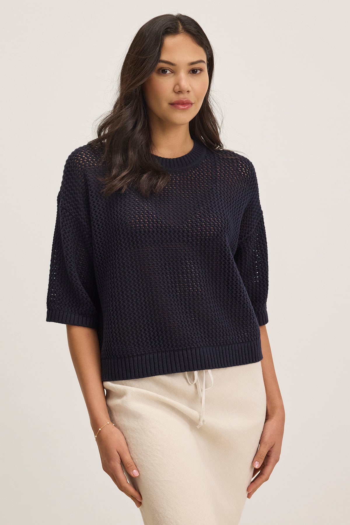 A woman stands wearing a relaxed fit, black VIENNA COTTON CASHMERE MESH SWEATER from Velvet by Graham & Spencer and a beige skirt, looking forward with a neutral expression, hands by her sides.