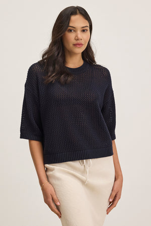 A woman stands wearing a relaxed fit, black VIENNA COTTON CASHMERE MESH SWEATER from Velvet by Graham & Spencer and a beige skirt, looking forward with a neutral expression, hands by her sides.