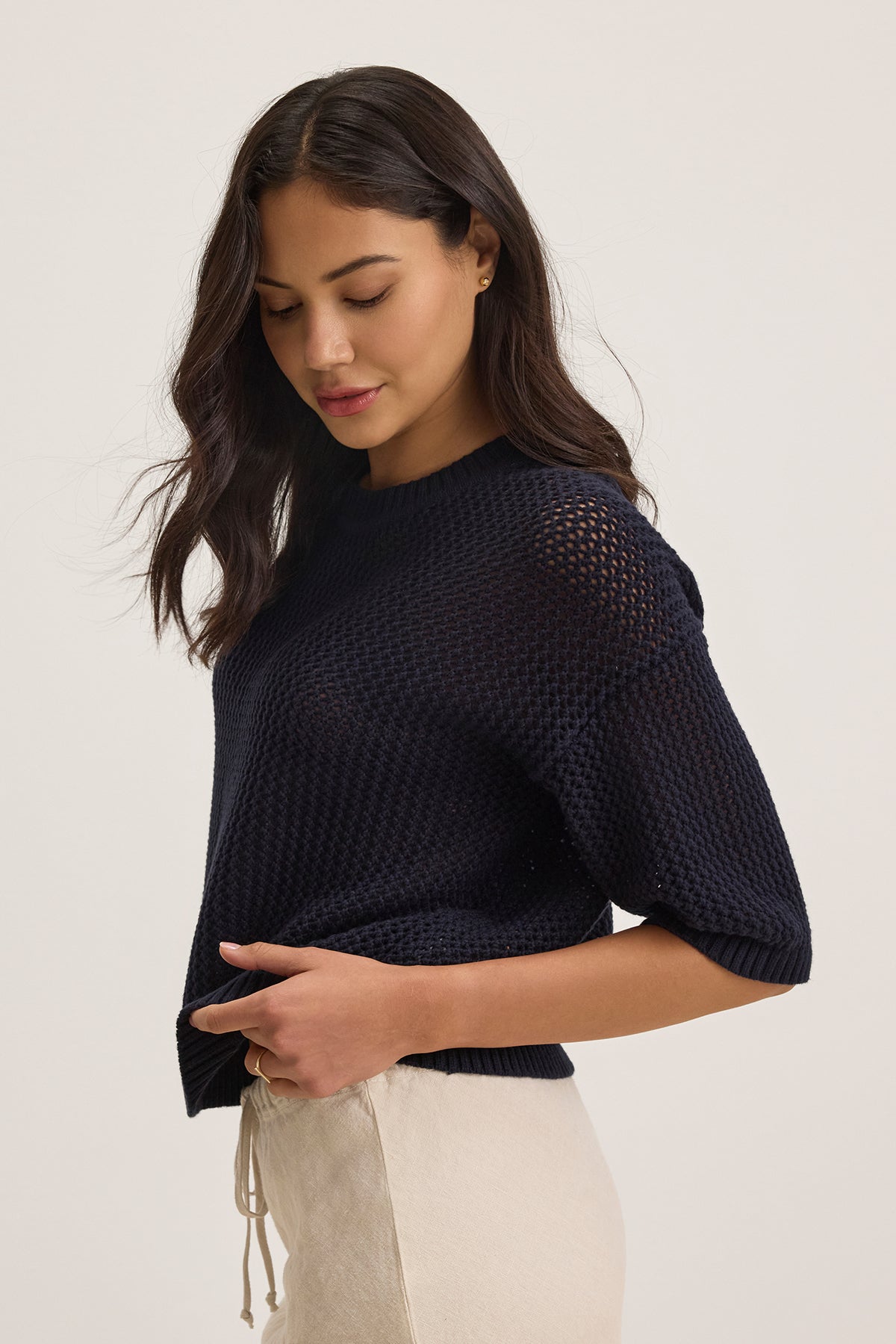 A woman in a relaxed-fit navy VIENNA COTTON CASHMERE MESH SWEATER by Velvet by Graham & Spencer and beige pants looks down against a light background.-38663172620481