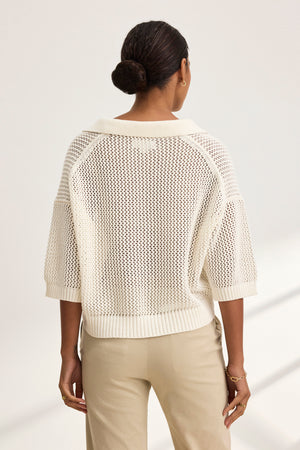 With hair in a bun, they stand facing away, dressed in a YORK COTTON CASHMERE MESH SWEATER POLO by Velvet by Jenny Graham. Their beige pants enhance the attire's soft elegance.