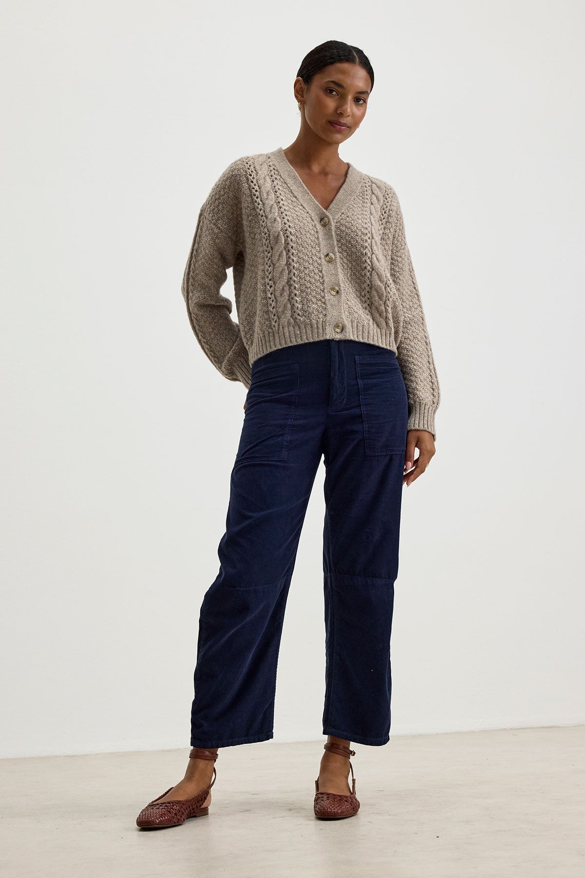Person standing against a plain background wearing an IZZY CARDIGAN by Velvet by Graham & Spencer with a cable knit pattern, navy blue wide-leg pants, and brown shoes.-37856185352385