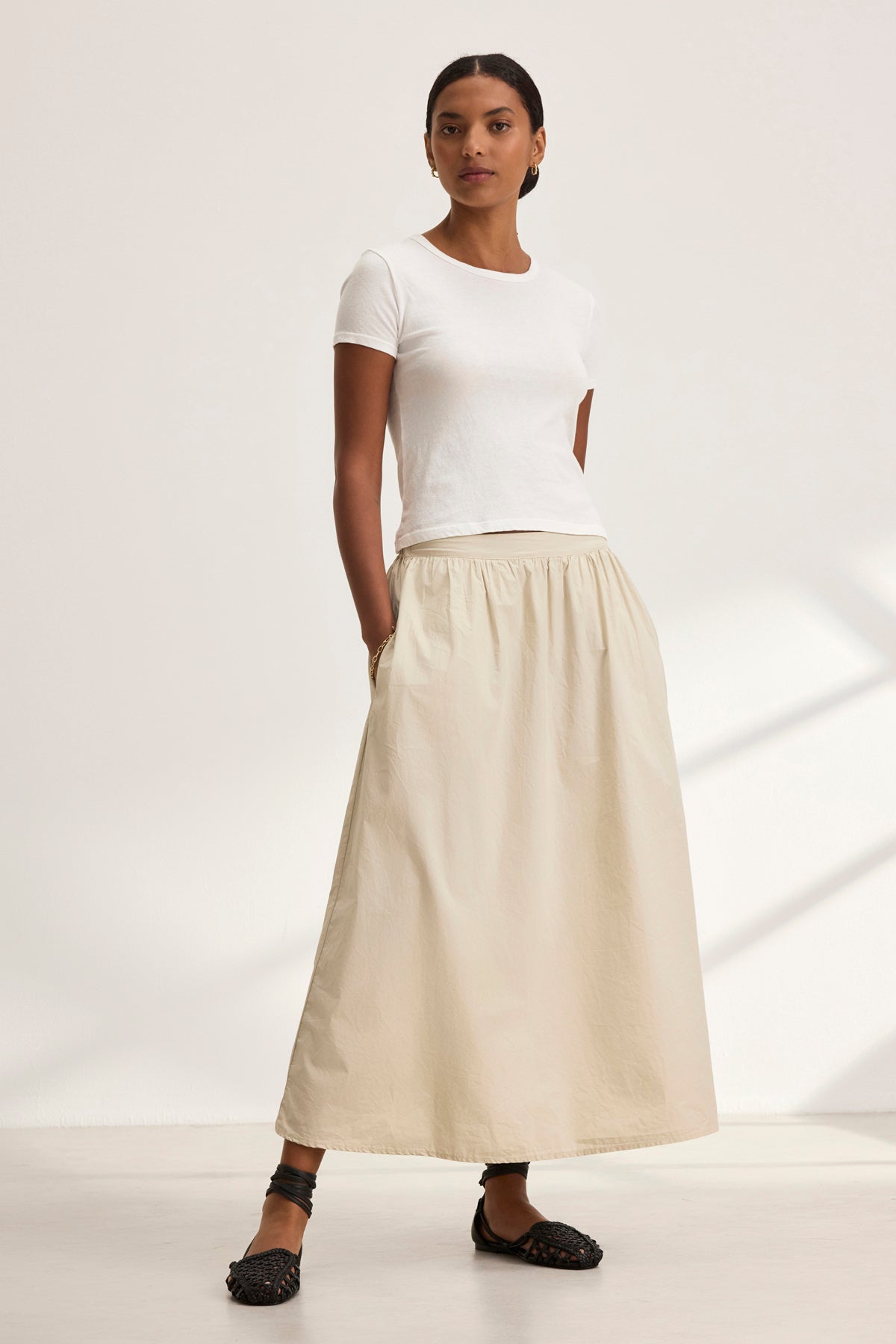 A person stands wearing a white t-shirt and the BELLA SKIRT, a long beige A-line skirt by Velvet by Graham & Spencer, against a plain background.-38957694714049