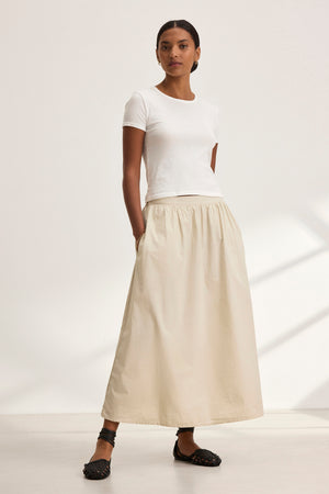 A person stands wearing a white t-shirt and the BELLA SKIRT, a long beige A-line skirt by Velvet by Graham & Spencer, against a plain background.