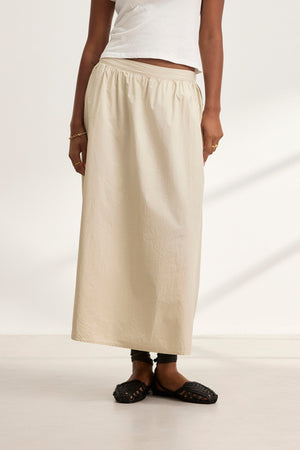 A person stands against a plain background wearing a white top, the BELLA SKIRT by Velvet by Graham & Spencer, and black woven flats.