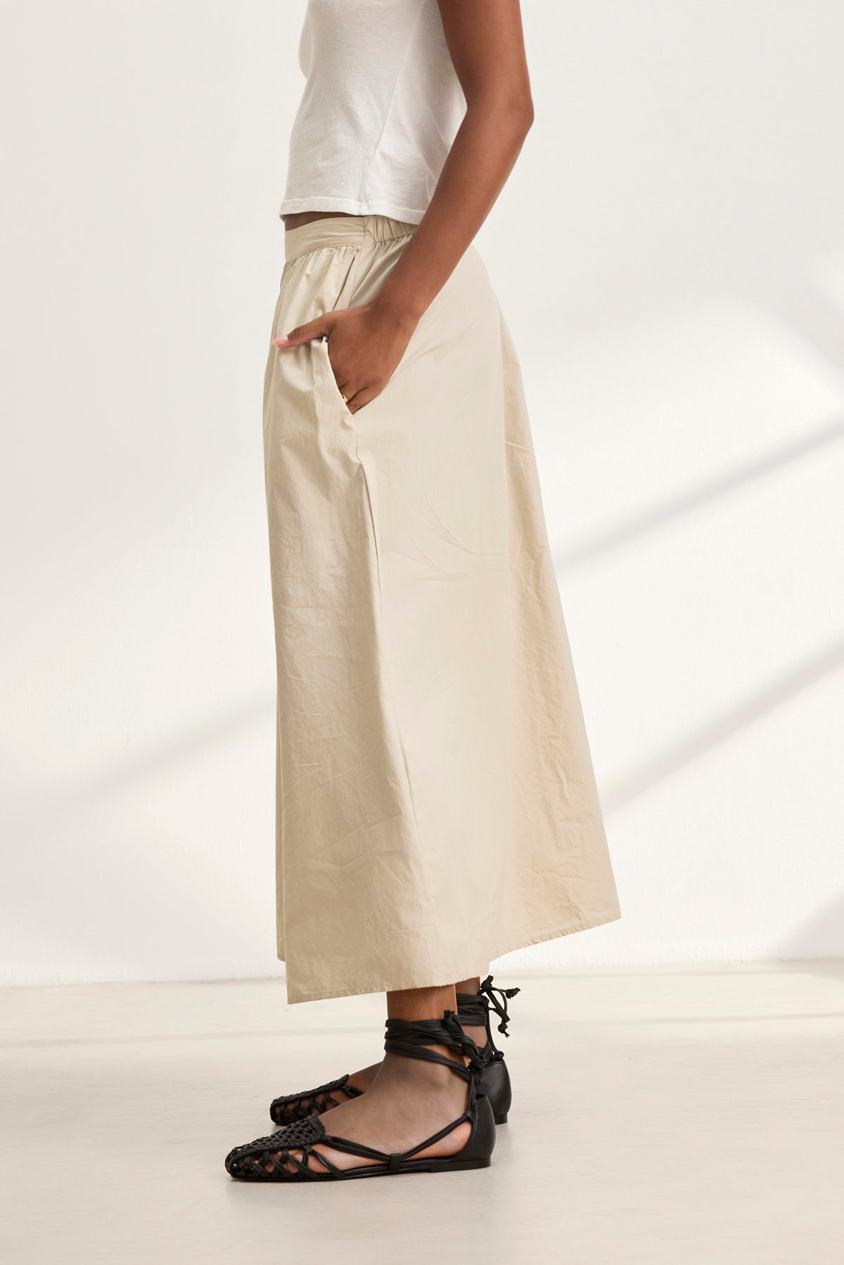 A person in a white top, beige wide-leg pants, and black lace-up ballet flats stands sideways with hands in pockets, exuding the effortless style of the BELLA SKIRT by Velvet by Graham & Spencer.-38957694648513