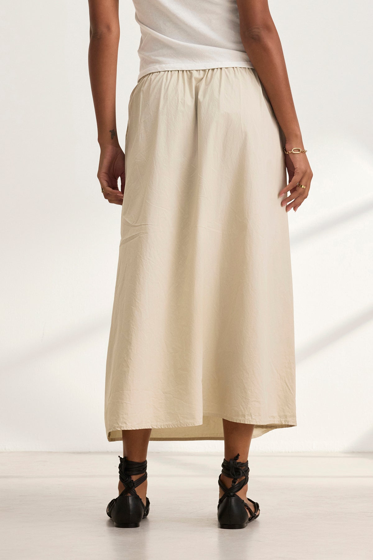 A person gracefully stands on a white floor, wearing a white top and the elegant BELLA SKIRT in beige by Velvet by Graham & Spencer, paired with black sandals, facing away.