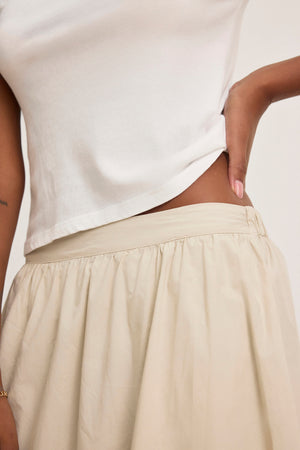 A person in a white top and the BELLA SKIRT by Velvet by Graham & Spencer, which is beige, made of cotton poplin with a midi length and elegant A-line silhouette, with one hand on their hip.
