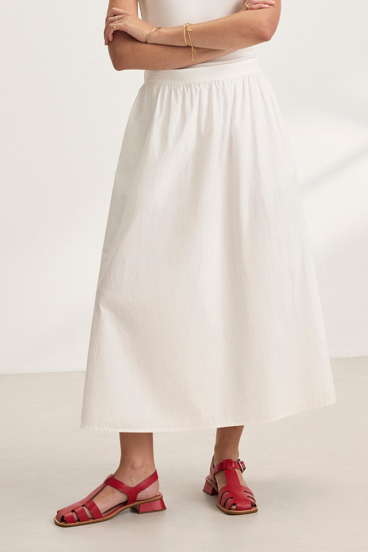 A person wearing the Velvet by Graham & Spencer BELLA SKIRT, a white cotton poplin A-line silhouette, paired with a tank top and red sandals, stands with arms crossed.