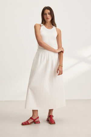 A woman in a white sleeveless top and Velvet by Graham & Spencer's BELLA SKIRT, an A-line midi-length cotton poplin skirt, paired with red sandals, stands against a plain white background, exuding elegance.