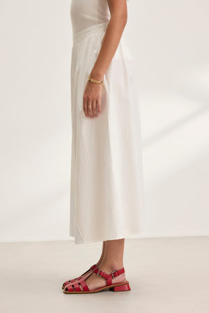 A person in a white BELLA SKIRT by Velvet by Graham & Spencer with an A-line cut and red sandals stands sideways in a well-lit room.