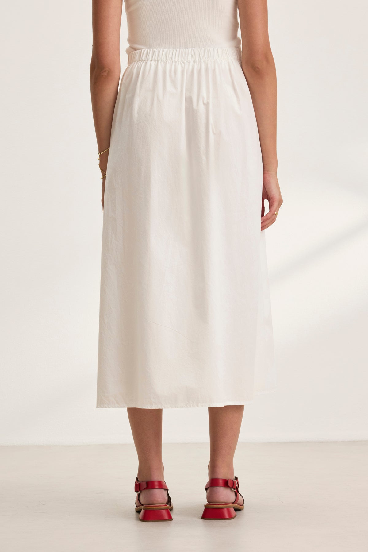 Back view of a person wearing Velvet by Graham & Spencer’s BELLA SKIRT—a plain white cotton poplin A-line skirt—and red sandals on a neutral background.-38802047140033