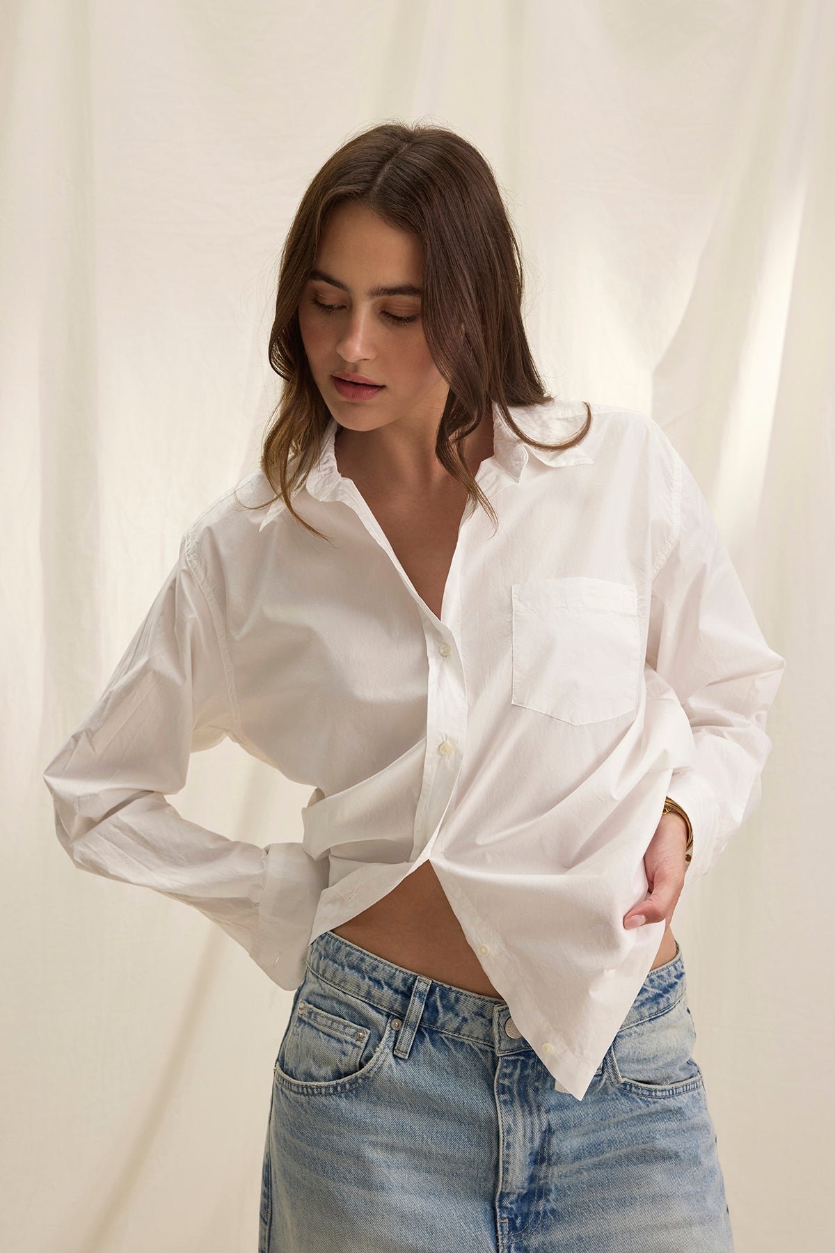 A woman wears the GRACE SHIRT by Velvet by Graham & Spencer—an oversized button-up in lightweight cotton poplin—paired with light blue jeans, standing against a plain, light-colored backdrop.-39273970958529