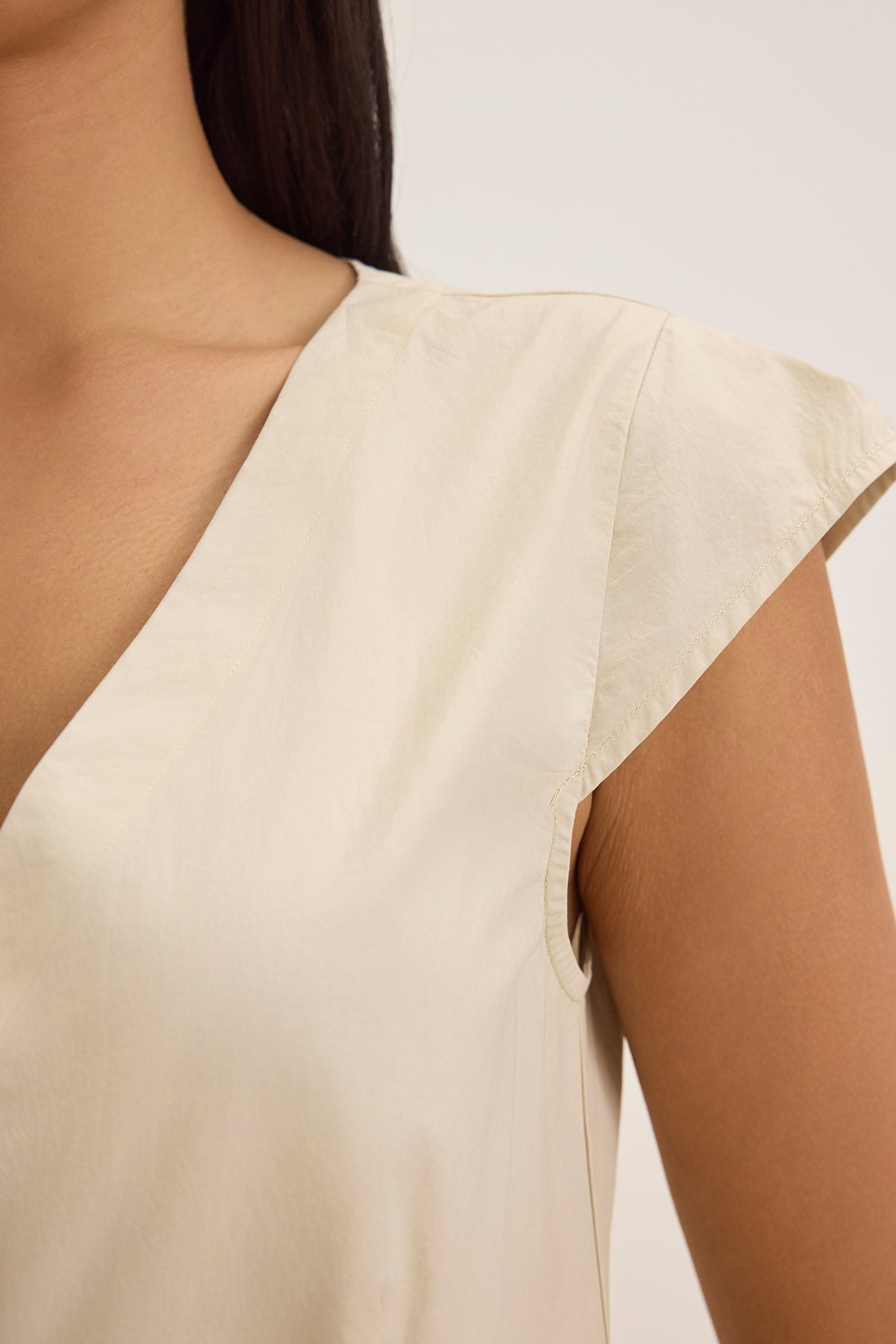 A close-up shows someone in the INETTE DRESS by Velvet by Graham & Spencer, a sleeveless V-neck beige top made of cotton poplin, highlighting the shoulder and upper chest against a minimal background.-38662805782721