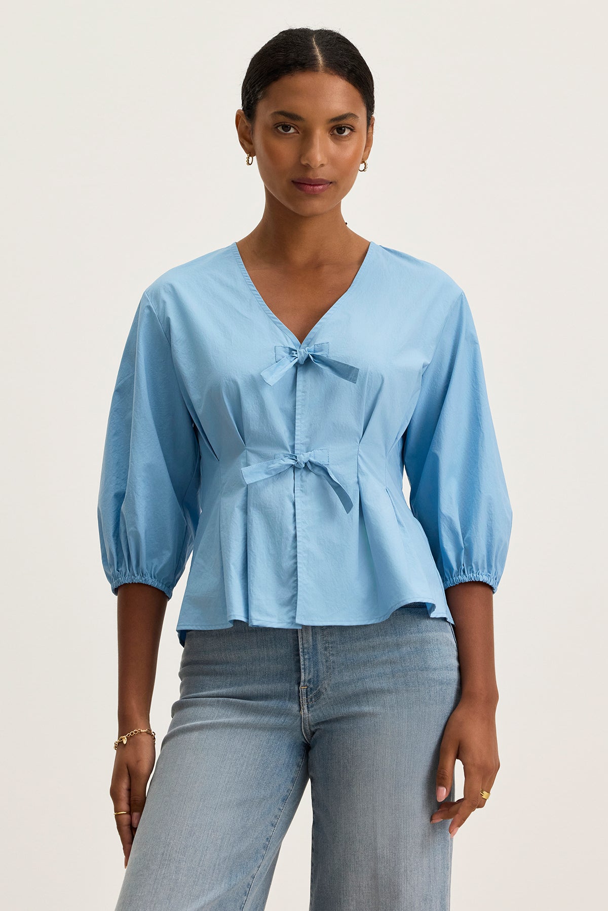 A woman wears the RIRI TOP by Velvet by Graham & Spencer—a relaxed fit, light blue cotton poplin blouse with front bows and 3/4 sleeves—paired with light blue jeans against a plain background.