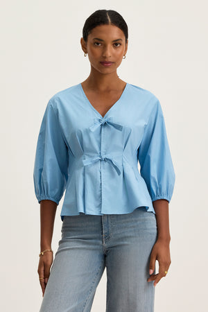 A woman wears the RIRI TOP by Velvet by Graham & Spencer—a relaxed fit, light blue cotton poplin blouse with front bows and 3/4 sleeves—paired with light blue jeans against a plain background.