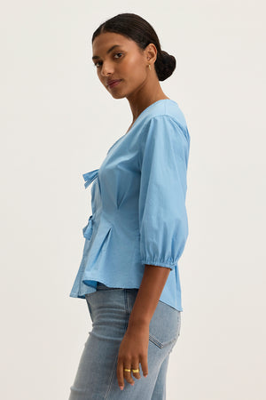 A woman stands in profile wearing the Velvet by Graham & Spencer RIRI TOP, a light blue cotton poplin blouse with relaxed fit and three-quarter sleeves, paired with light blue jeans against a plain background.