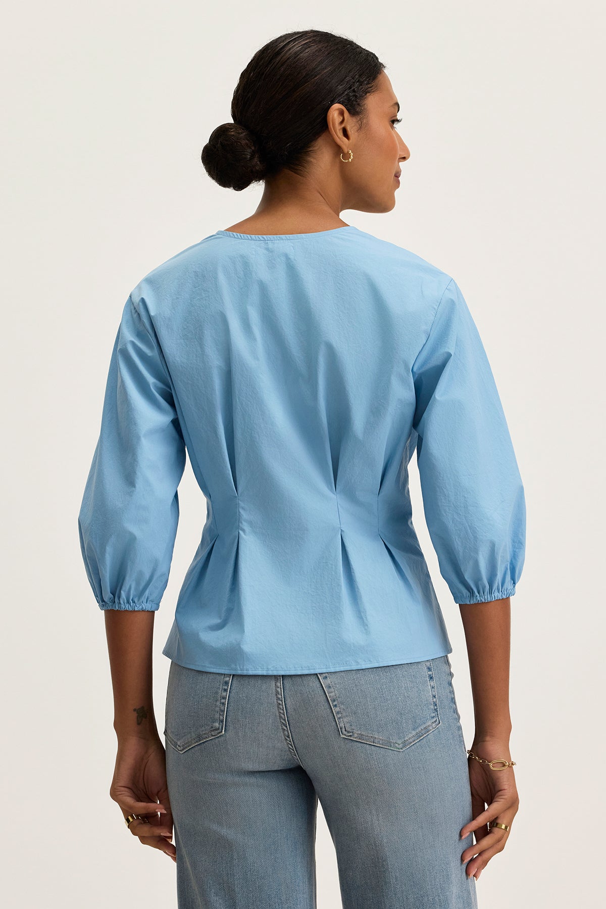 A woman faces away, wearing the Velvet by Graham & Spencer RIRI TOP—a relaxed fit, light blue three-quarter sleeve blouse—with light blue jeans, shown against a plain light background.