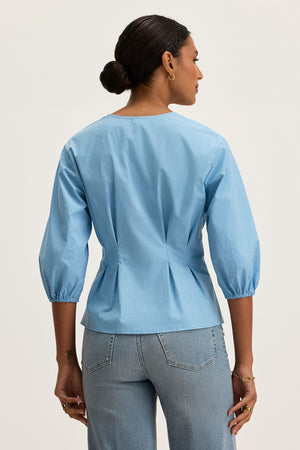 A woman faces away, wearing the Velvet by Graham & Spencer RIRI TOP—a relaxed fit, light blue three-quarter sleeve blouse—with light blue jeans, shown against a plain light background.