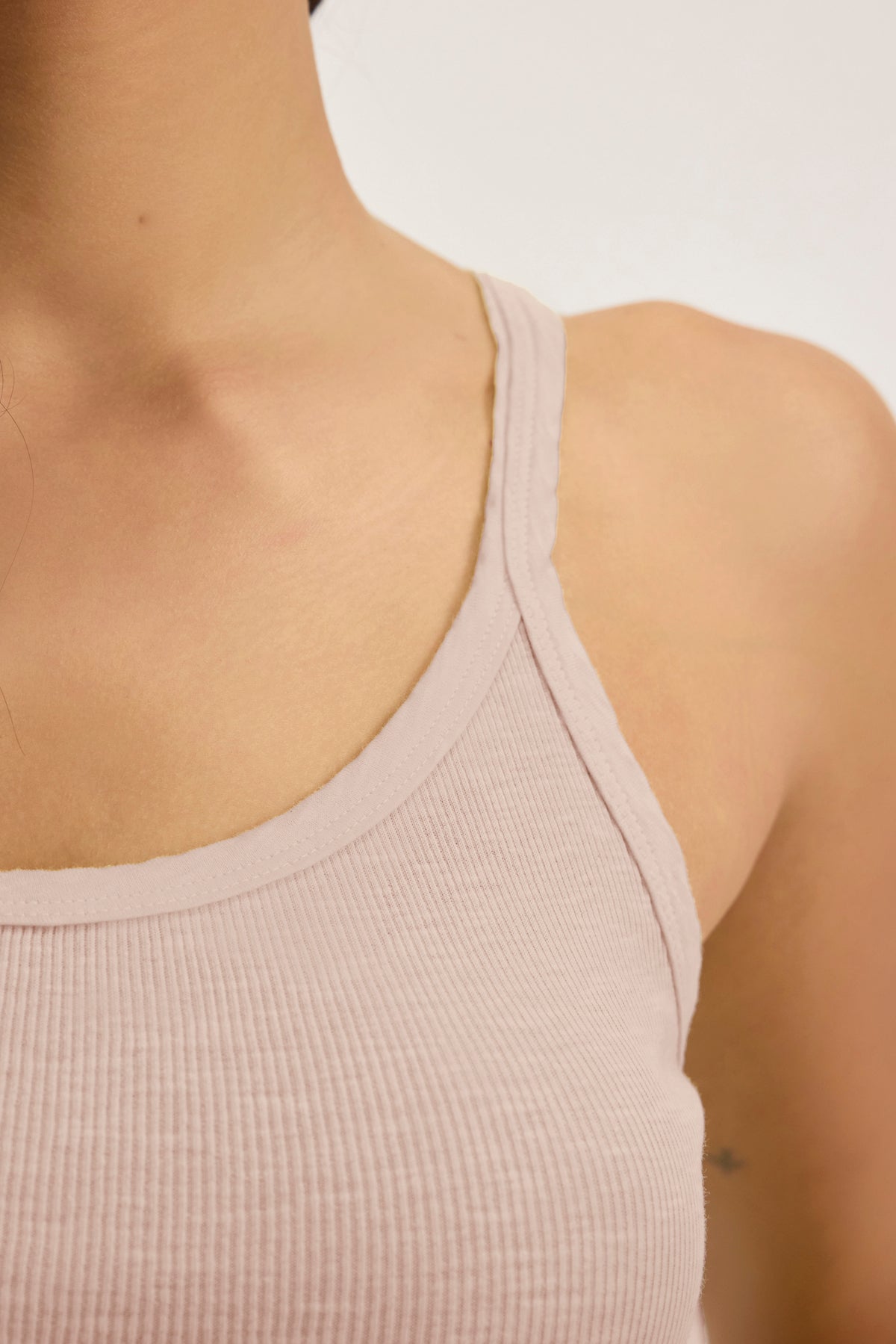 A close-up shows someone wearing the ALIZA Ribbed Cotton Layering Tank by Velvet by Graham & Spencer in light pink, revealing their shoulder and collarbone against a plain background, ideal for layering.-38928214524097