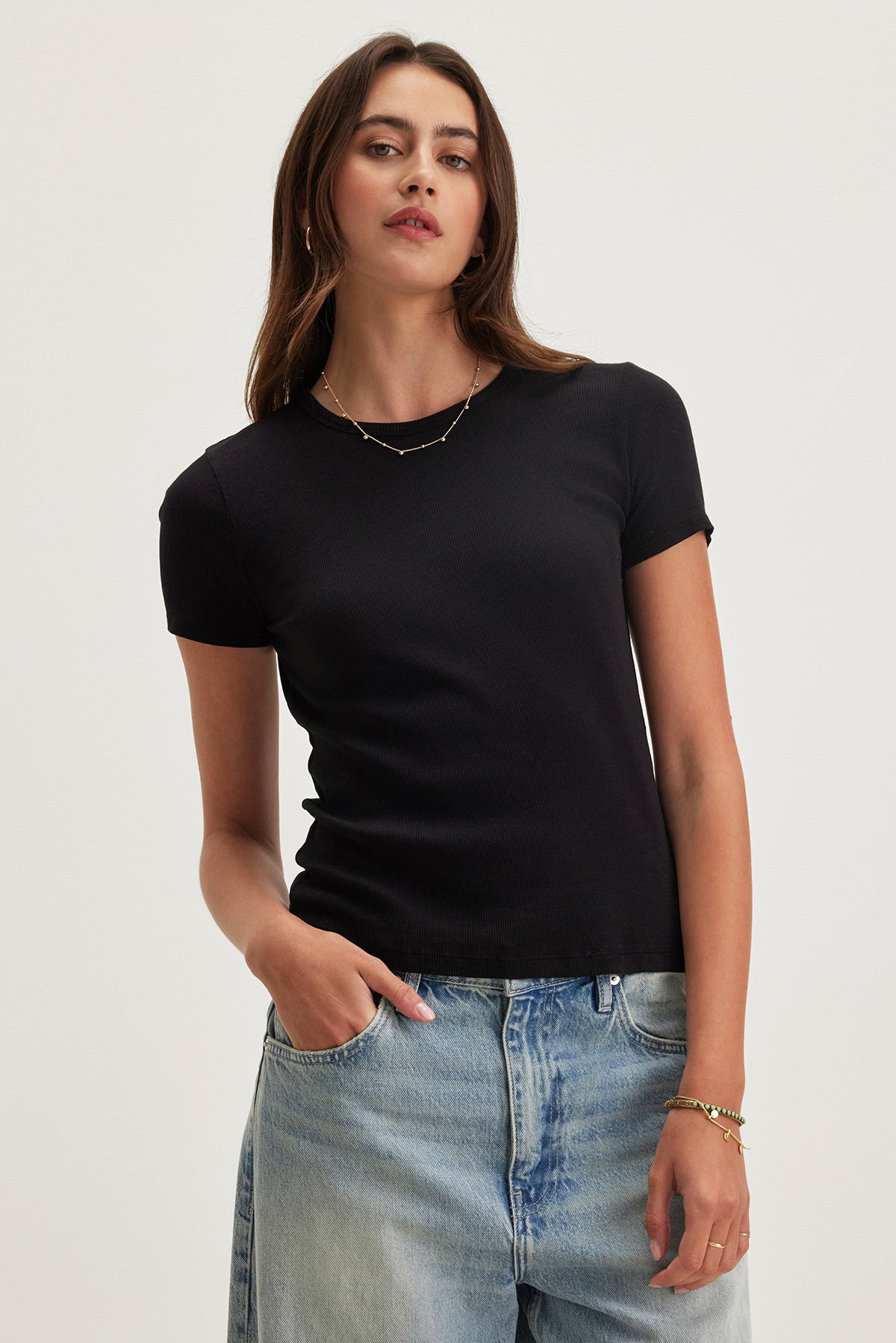 A person stands against a plain background wearing the BRENNY TEE by Velvet, featuring a ribbed cotton texture and crew neckline, paired with light blue jeans. The tee complements their necklace and bracelet.
