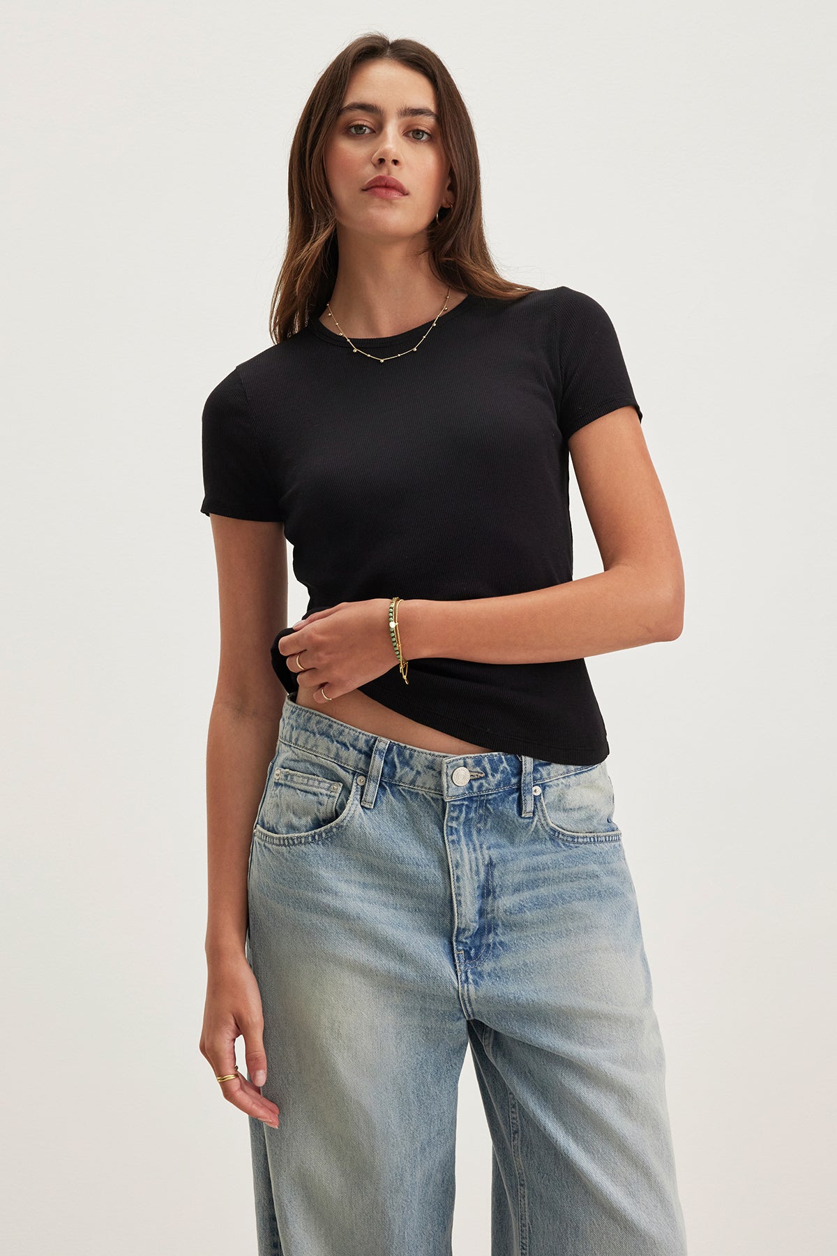 A model wears the BRENNY TEE, a black ribbed cotton shirt by Velvet by Graham & Spencer, paired with blue jeans. They stand against a neutral background, highlighting its relaxed crew neckline.