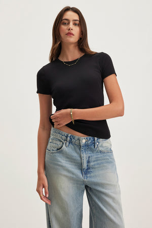 A model wears the BRENNY TEE, a black ribbed cotton shirt by Velvet by Graham & Spencer, paired with blue jeans. They stand against a neutral background, highlighting its relaxed crew neckline.