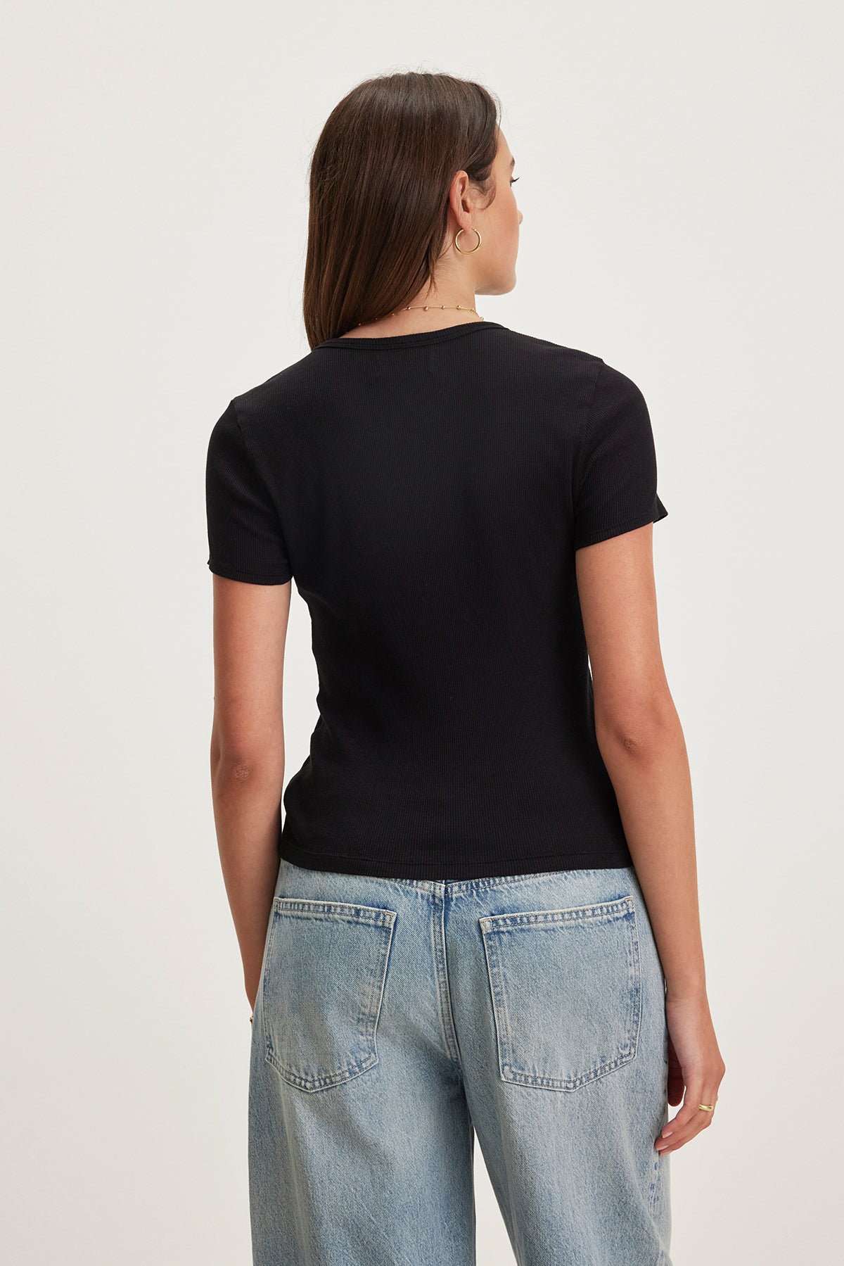 A woman with long hair, wearing the BRENNY TEE by Velvet by Graham & Spencer—a black cotton slub knit t-shirt with a stylish crew neckline and cropped silhouette—and light blue jeans, faces away from the camera against a plain background.