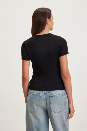 A woman with long hair, wearing the BRENNY TEE by Velvet by Graham & Spencer—a black cotton slub knit t-shirt with a stylish crew neckline and cropped silhouette—and light blue jeans, faces away from the camera against a plain background.