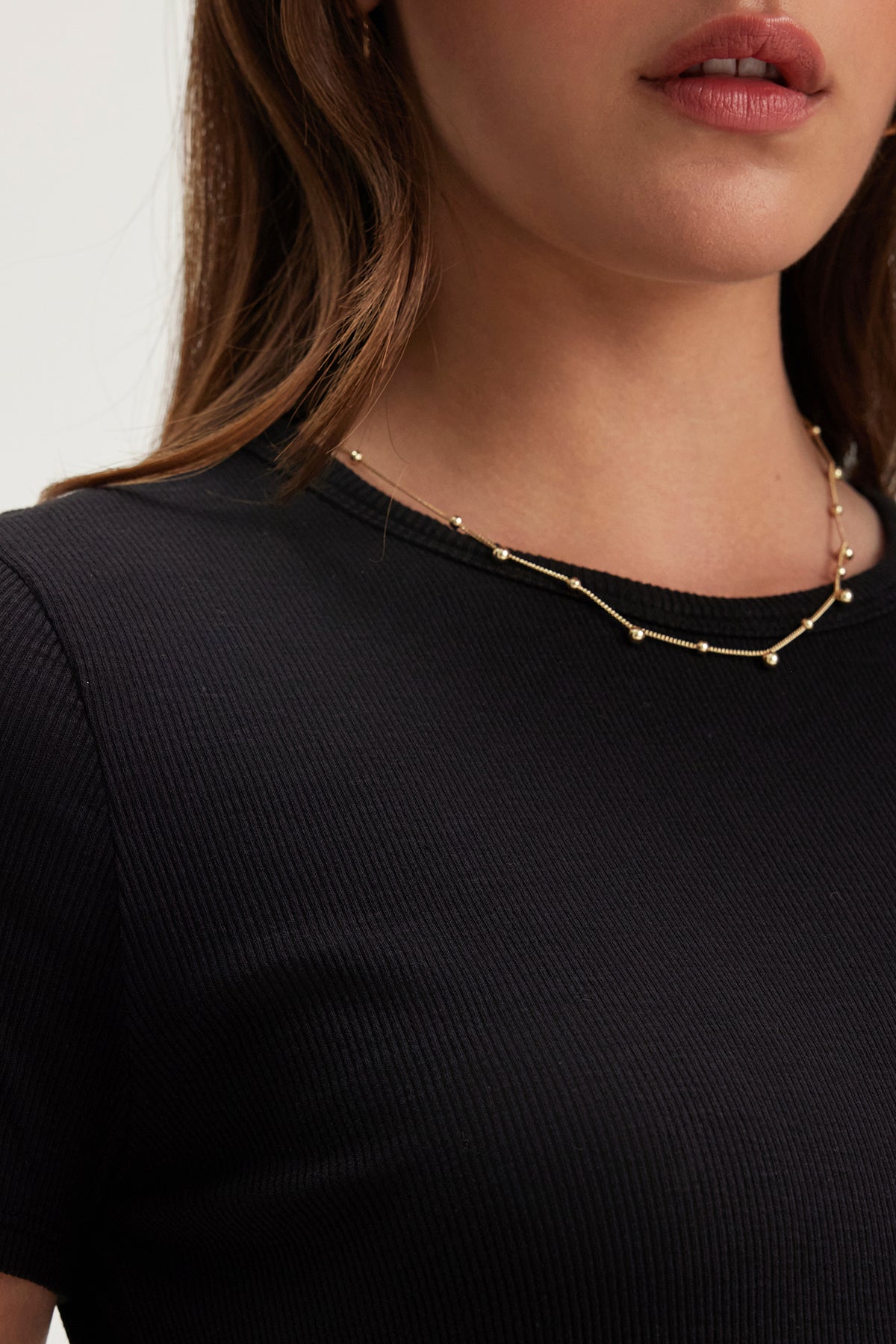 A person wearing the BRENNY TEE, a black ribbed cotton knit top with a crew neckline by Velvet by Graham & Spencer, paired with a delicate gold necklace featuring small metallic beads.-38662911426753