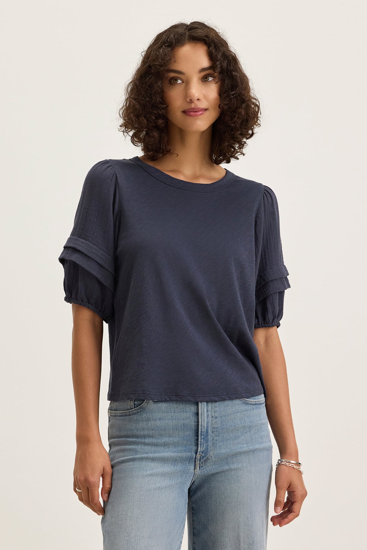 A person with short curly hair wears the HADEN TOP by Velvet by Graham & Spencer in navy blue, featuring puff sleeves and a relaxed fit, paired with light blue jeans, standing against a plain light background.