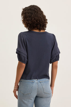 A person with curly hair is seen from the back wearing the HADEN TOP by Velvet by Graham & Spencer, a navy cotton slub short-sleeve top with puff sleeves, paired with light blue jeans against a plain background.