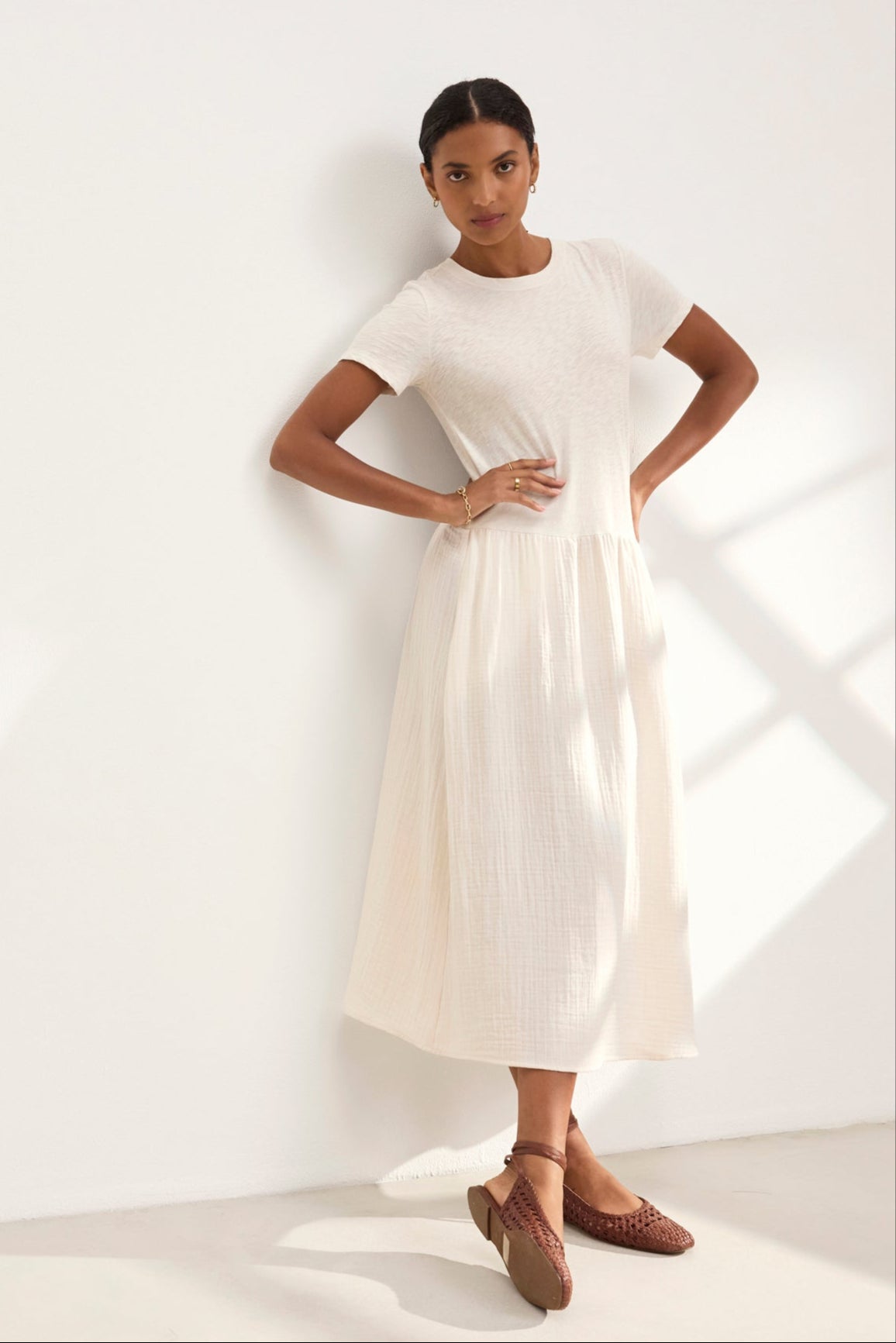 A woman in the ITZA DRESS by Velvet by Graham & Spencer leans against a wall in soft lighting, wearing brown woven shoes.-38918714458305