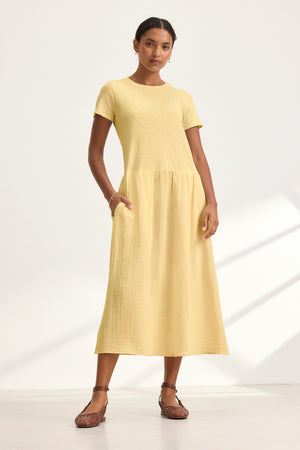 A woman stands against a light background wearing the Velvet by Graham & Spencer ITZA DRESS in yellow with side seam pockets, paired with brown woven shoes.