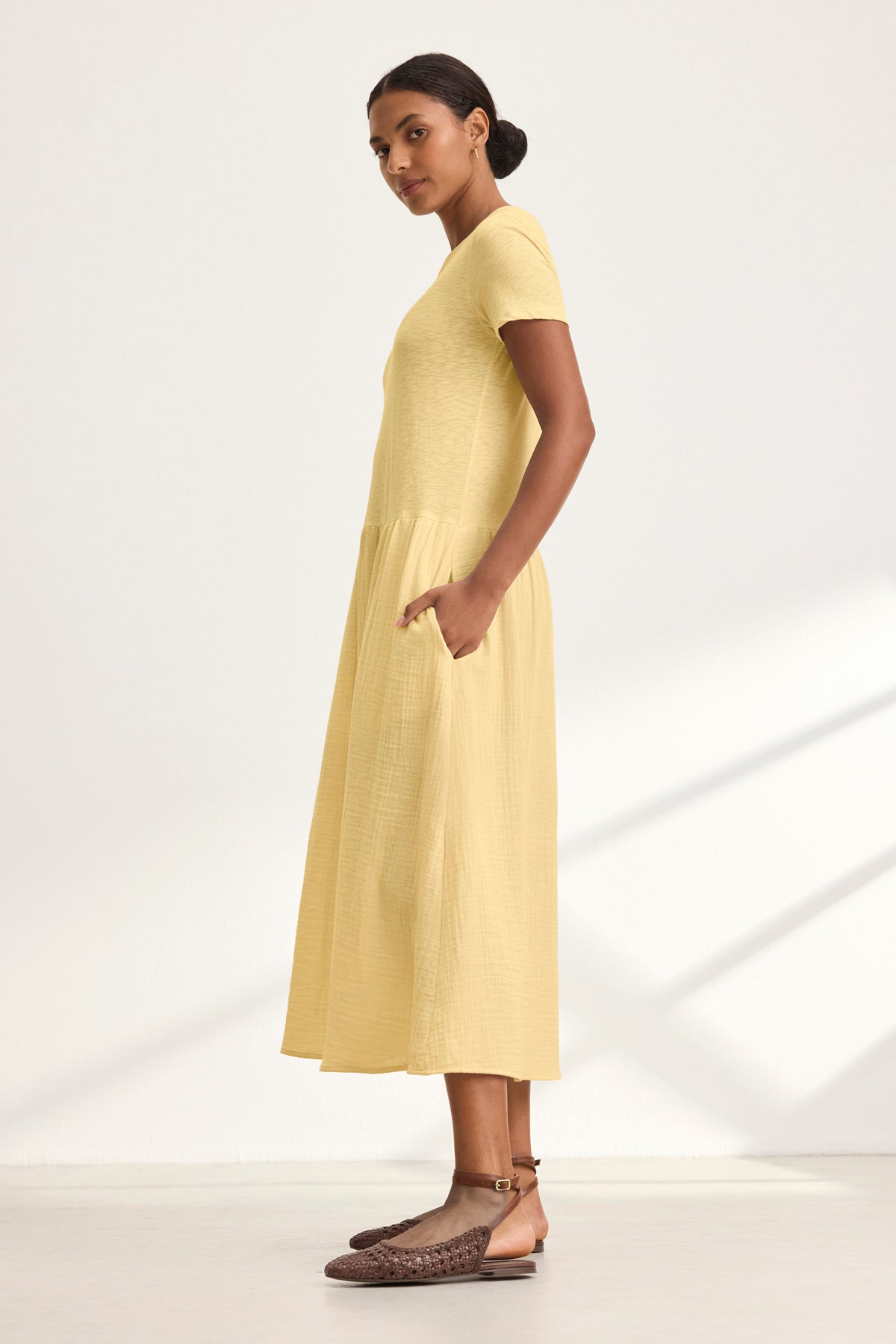 A woman stands in profile wearing the light yellow ITZA DRESS by Velvet by Graham & Spencer, a cotton gauze midi with side seam pockets, paired with brown woven flat sandals against a plain light background.