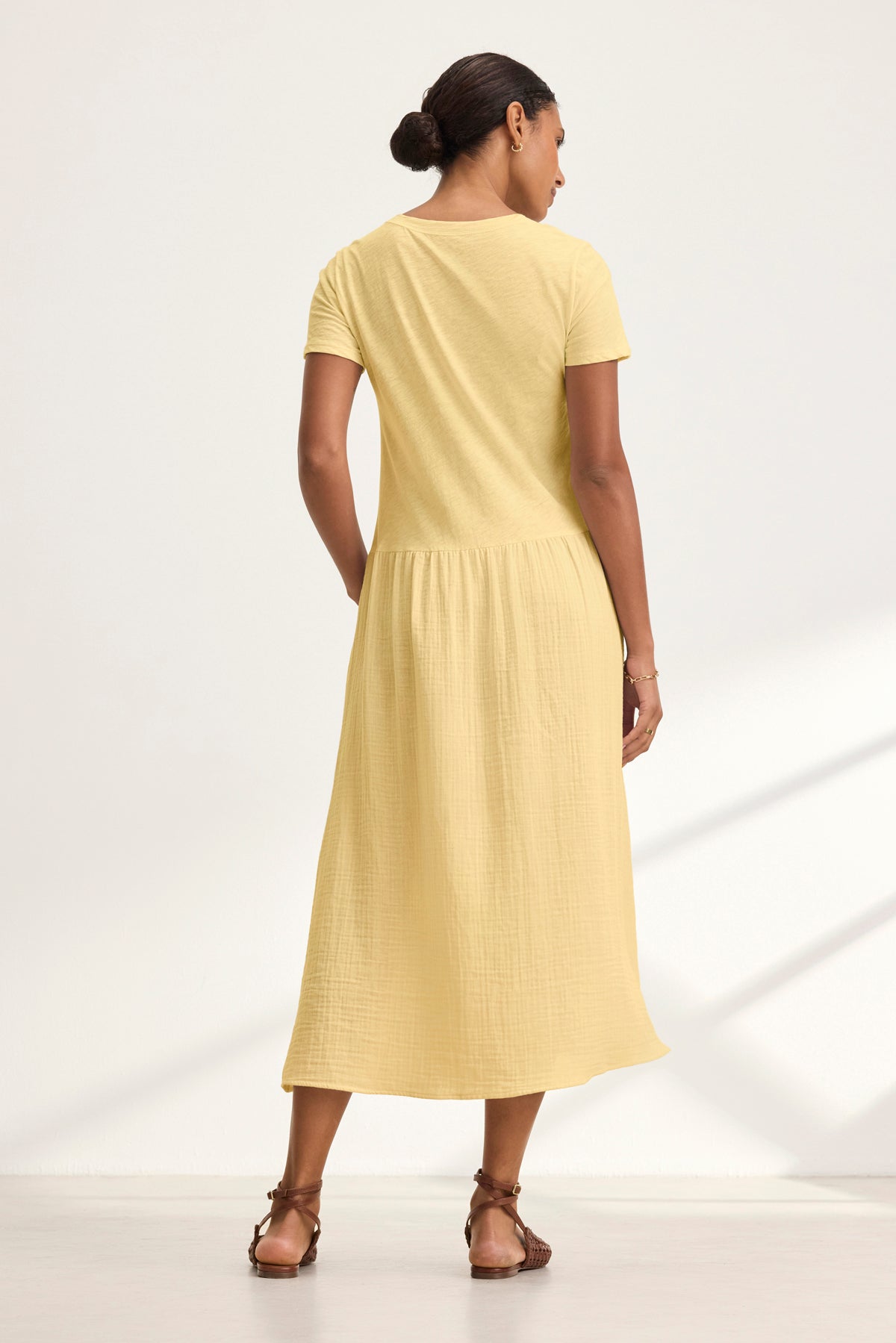 A person stands facing away, wearing the ITZA DRESS by Velvet by Graham & Spencer—a short-sleeved, light yellow midi dress with side seam pockets—paired with brown sandals and hair tied back, against a plain light background.
