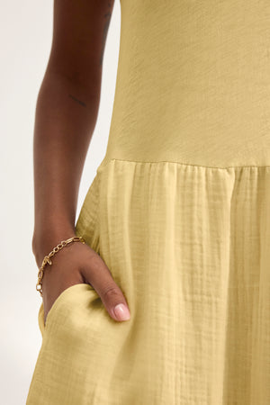 Close-up of a person wearing the ITZA DRESS by Velvet by Graham & Spencer—a light yellow midi dress with a gathered waist, crew neckline, pocket detail, and accessorized with a gold bracelet.