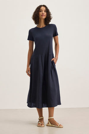 A woman stands indoors against a plain background, wearing the ITZA DRESS by Velvet by Graham & Spencer—a navy midi dress with a crew neckline, short sleeves, and a gathered waist—paired with beige sandals.