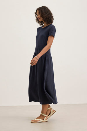 A woman with curly hair models the ITZA DRESS by Velvet by Graham & Spencer—a loose, short-sleeve navy midi dress with a crew neckline—paired with beige sandals, standing against a plain light background.