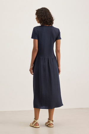 A woman faces away, wearing the ITZA DRESS by Velvet by Graham & Spencer—a navy blue midi dress with a crew neckline—paired with beige sandals against a plain light background.