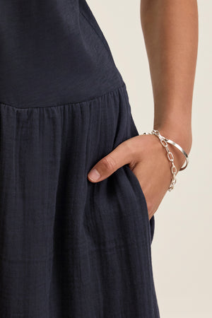A person wearing the ITZA DRESS by Velvet by Graham & Spencer, a dark blue midi with a crew neckline, their right hand in the pocket, styled with a silver chain bracelet and bangle.