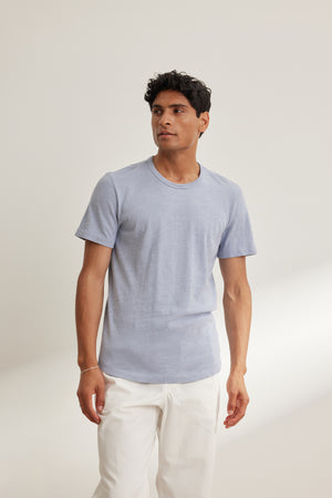 A man wearing the SPADER TEE by Velvet by Graham & Spencer, a relaxed fit light blue Peruvian cotton crewneck, and white pants stands against a plain light background.
