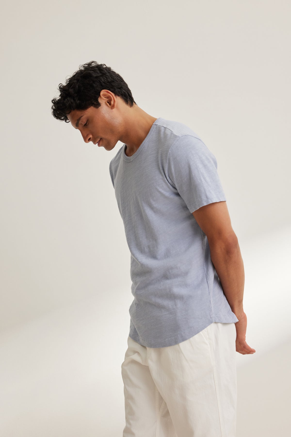 A man wears the SPADER TEE by Velvet by Graham & Spencer—a relaxed fit, light blue Peruvian cotton crewneck—paired with white pants, standing against a plain light background, looking down with hands behind his back.